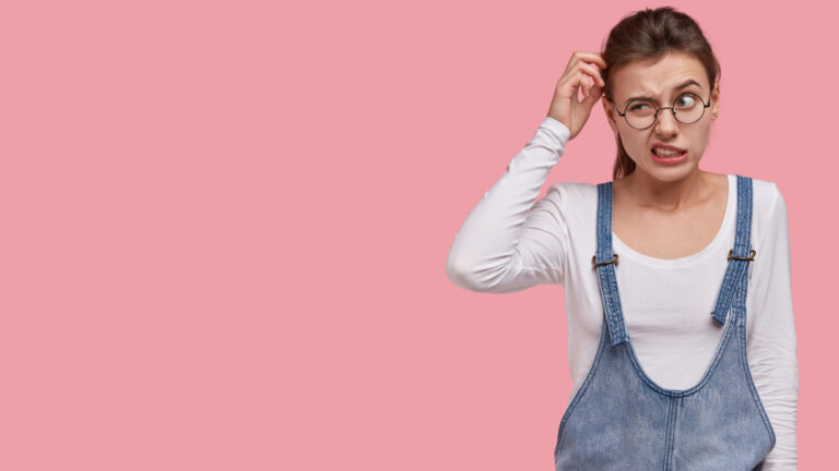 Photo of puzzled woman scratches head in bewilderment, thinks about finding right solution, wears trendy sarafan, round spectacles, models over pink background with free space for information