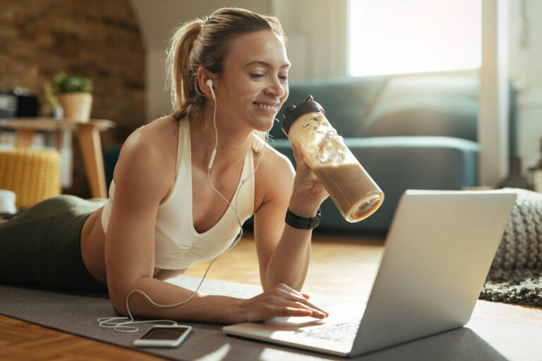 Happy athletic woman drinking protein shake while using laptop o