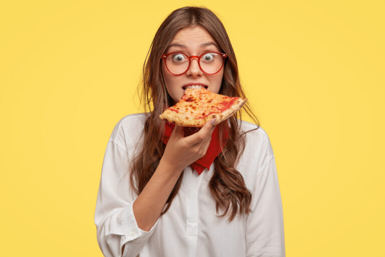 Emotive beautiful lady bites delicious pizza, looks directly at camera, has time for snack, visits pizzeria, surprised with low prices, models over yellow background
