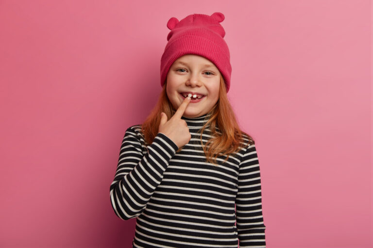 Pretty small ginger girl shows her first adult two teeth, laughs and rejoices, expresses positive emotions, keeps mouth opened, prepares for oral checkup, dressed in striped sweater and pink hat